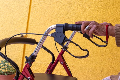 H&auml;nde einer alten Frau halten die Griffe eines Rollators. Im Hintergrund eine gelb gestrichene Wand. 