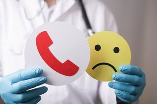 Ein Arzt mit blauen Handschugen und einem weißen Kittel hält einen traurigen Smiley aus Pappe in der einen Hand, in der anderen ein rotes Telefonhörer-Symbol