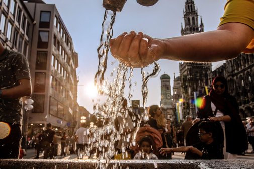 Blick in eine Innenstadt mit H&auml;usern und Menschen, im Vordergrund eine Hand, die Wasser aus einem Hahn oder Brunnen sch&ouml;pft.