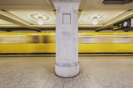 Die U-Bahnlinie 3 verkehrt am Ruedesheimer Platz in Wilmersdorf in Berlin. Man sieht die U-Bahn verschwommen, sie fährt gerade ein. Ein Stützpfeiler teilt das Foto in zwei Hälften.