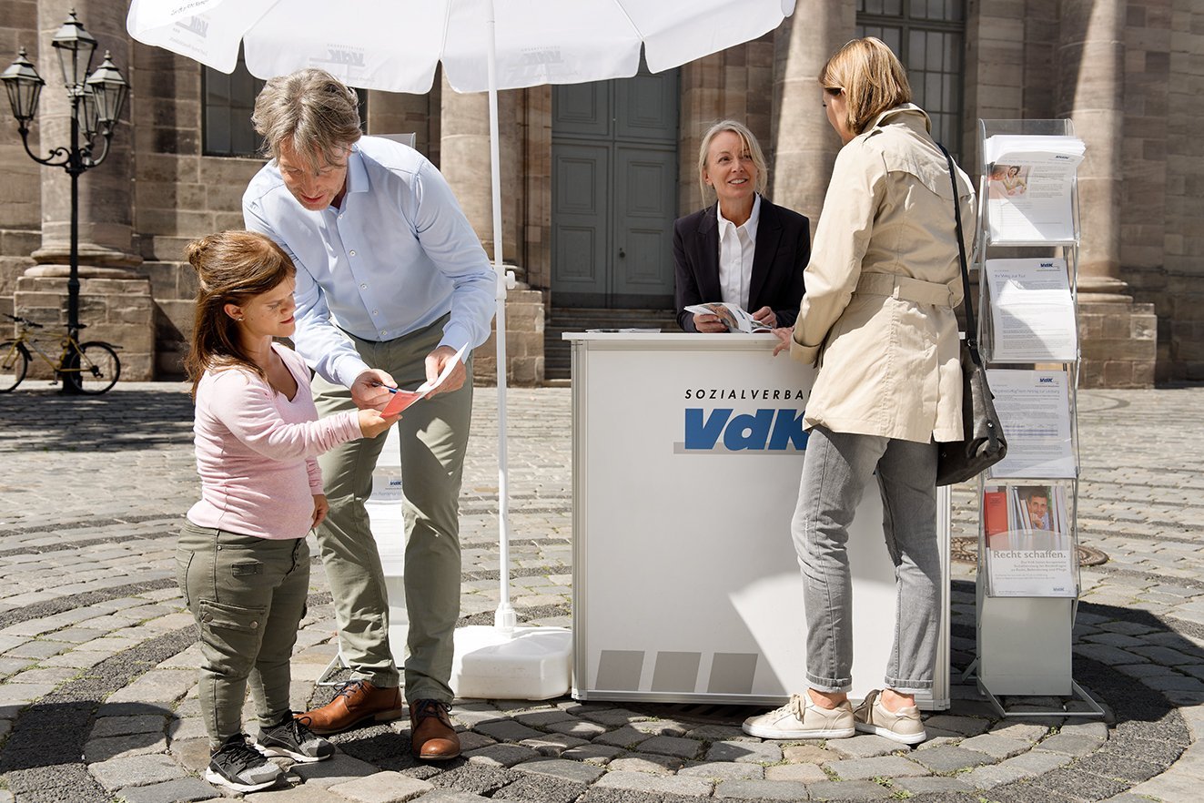 Ein VdK-Infostand mit einer Mitarbeiterin, davor verschiedene Menschen, die sich informieren wollen.