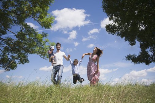 Eine junge Familie mit zwei kleineren Kindern auf einer Wiese vor blauem Himmel, der Vater tr&auml;gt ein Kind auf dem Arm, das andere Kind halten die Eltern zwischen sich und lassen es "fliegen".