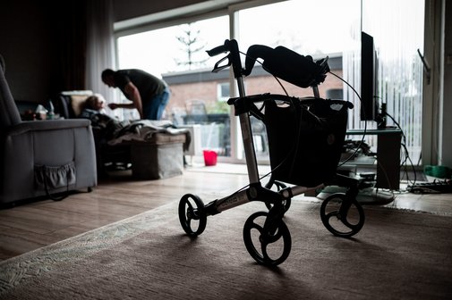 Blick in ein dunkles Wohnzimmer. Im Vordergrund steht ein Rollator. Im Hintergrund sieht man eine sitzende Person, die von einer anderen Person, die sich &uuml;ber sie beugt, gef&uuml;ttert wird. Symbolfoto f&uuml;r Angeh&ouml;rigenpflege