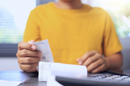 Eine Person im gelben Pullover sitzt an einem Schreibtisch, vor sich einen Taschenrechner und ein Dokument, wahrscheinlich eine Rechnung, als Symbol f&uuml;r steigende Kosten