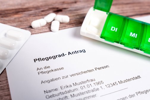Ein ausgedruckter Muster-Antrag auf einen Pflegegrad, daneben liegen Tabletten-Blister und eine Pillenbox