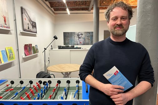 Benjamin Salzmann im Young Carer Zentrum in Berlin-Moabit, er steht an eine S&auml;ule gelehnt und h&auml;lt einen Flyer in den H&auml;nden, hinter ihm steht ein Kickertisch.