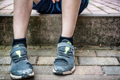 Ein Junge sitzt auf einem Bordstein, seine Turnschuhe sind abgenutzt und stellenweise kaputt.