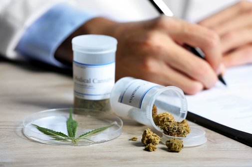 Ein Arzt schreibt etwas auf einen Block. Neben ihm auf dem Schreibtisch sind kleine Beh&auml;lter mit medizinischem Cannabis in Form von Cannabisbl&uuml;ten.