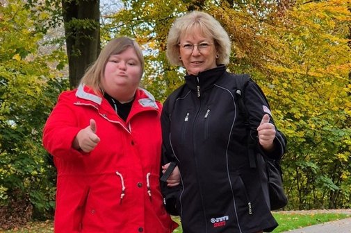 Zwei Frauen stehen auf einem Parkweg. Die Frau links trägt eine rote Jacke, braune Hose und schwarze Schuhe. Die Frau rechts trägt eine schwarze Jacke, rote Hose und graue Stiefeletten. Beide zeigen mit einer Hand den Daumen nach oben und lächeln. Im Hintergrund herbstlich gefärbte Bäume mit gelb-grünem Laub.