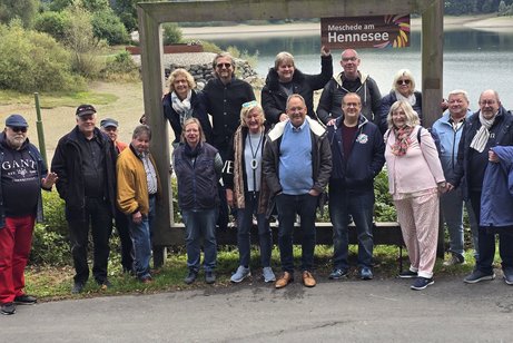 Gruppenbild mit Menschen, Frauen und Männern unterschiedlichen Alters. Im Hintergrund ein Gewässer.