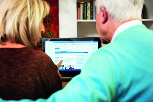 Eine Frau und ein Mann sitzen nebeneinander an einem Tisch und schauen auf einen Laptop-Bildschirm. Die Frau zeigt mit dem Finger auf die Webseite des Sozialverbands VdK Deutschland, die auf dem Bildschirm geöffnet ist. Der Bildschirm zeigt den Bereich „Ratgeber Recht“. Beide Personen sind von hinten zu sehen.