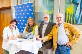 Zwei Frauen und zwei Männer stehen an einem Stehtisch und lachen in die Kamera.