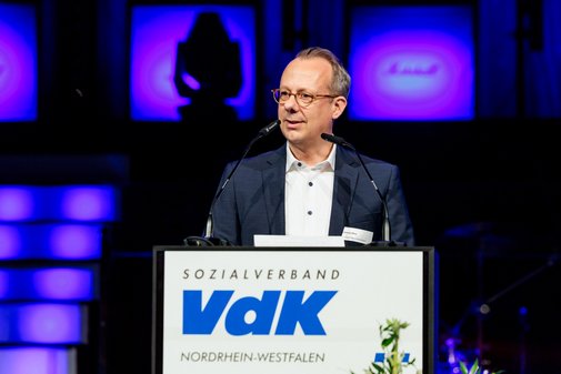 Carsten Ohm, VdK-Vorstand, steht an einem Pult mit der Aufschrift VdK. Er hat k&uuml;rzere Haare, tr&auml;gt eine Brille sowie ein blaues Sakko und ein wei&szlig;es Hemd.