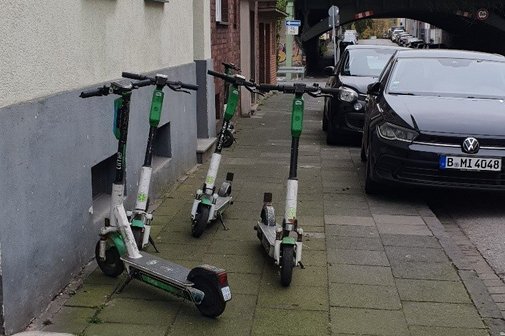 Mehrere E-Scooter sind mitten auf einem Bürgersteig abgestellt. Rechts auf der Gehwegkante parken Autos. Für Fußgänger bleibt nur eine kleine Lücke.