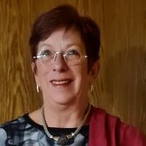 Portrait einer Frau mit kurzen rotbraunen Haaren, kleiner runder Brille und silbernen Ohrringen und Halskette vor einer holzvertäfelten braunen Wand.