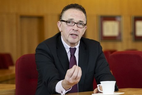 Prof. Dr. Landscheidt, ein Mann mit dunklem Kurzhaarschnitt und Brille, sitzt in Anzug und dunkelroter Kravatte an einem Tisch in einem holzvertäfelten Saal und gestikuliert energisch. 