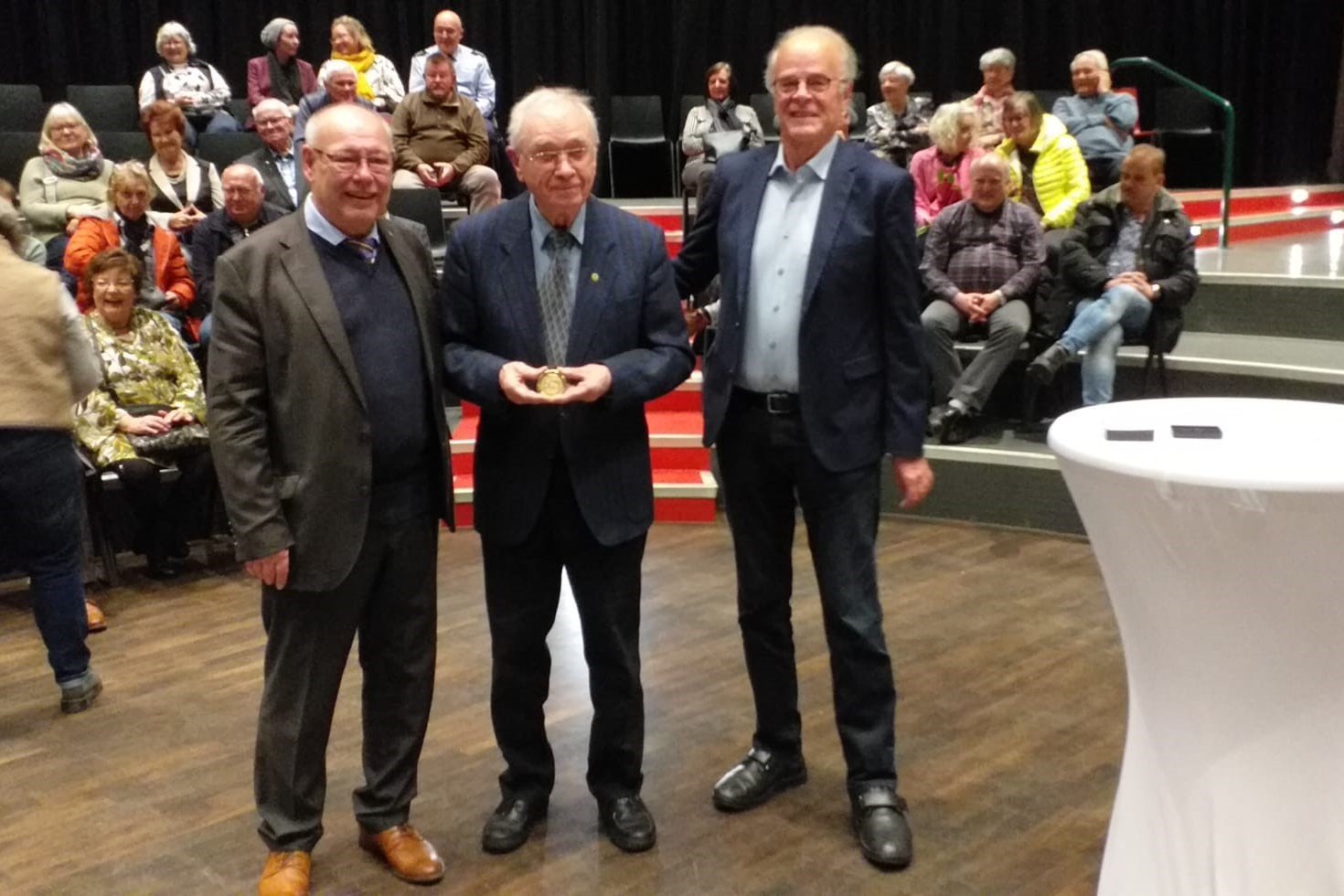 Drei Männer stehen nebeneinander in einem Saal. Rechts von ihnen ist ein Stehtisch mit weißer Husse, im Hintergrund viele Menschen.