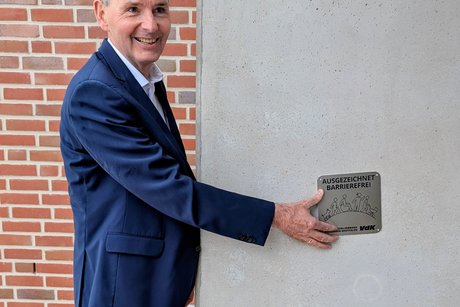 Ein Mann h&auml;lt die Barrierefrei-Plakette aus Edelstahl an eine graue Betonwand