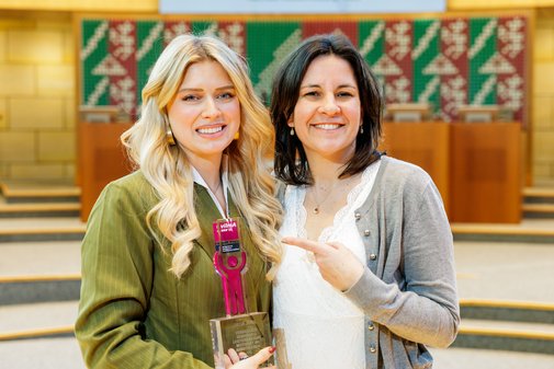 Eine Frau mit langen blonden Haaren und grüner Bluse hält den vilmA-Preis. Den Arm um ihre Schultern gelegt steht neben ihr eine Frau mit dunklen, mittellangen Haaren und weißer Bluse, mit dem Zeigefinder der freien Hand auf den Preis zeigend. 