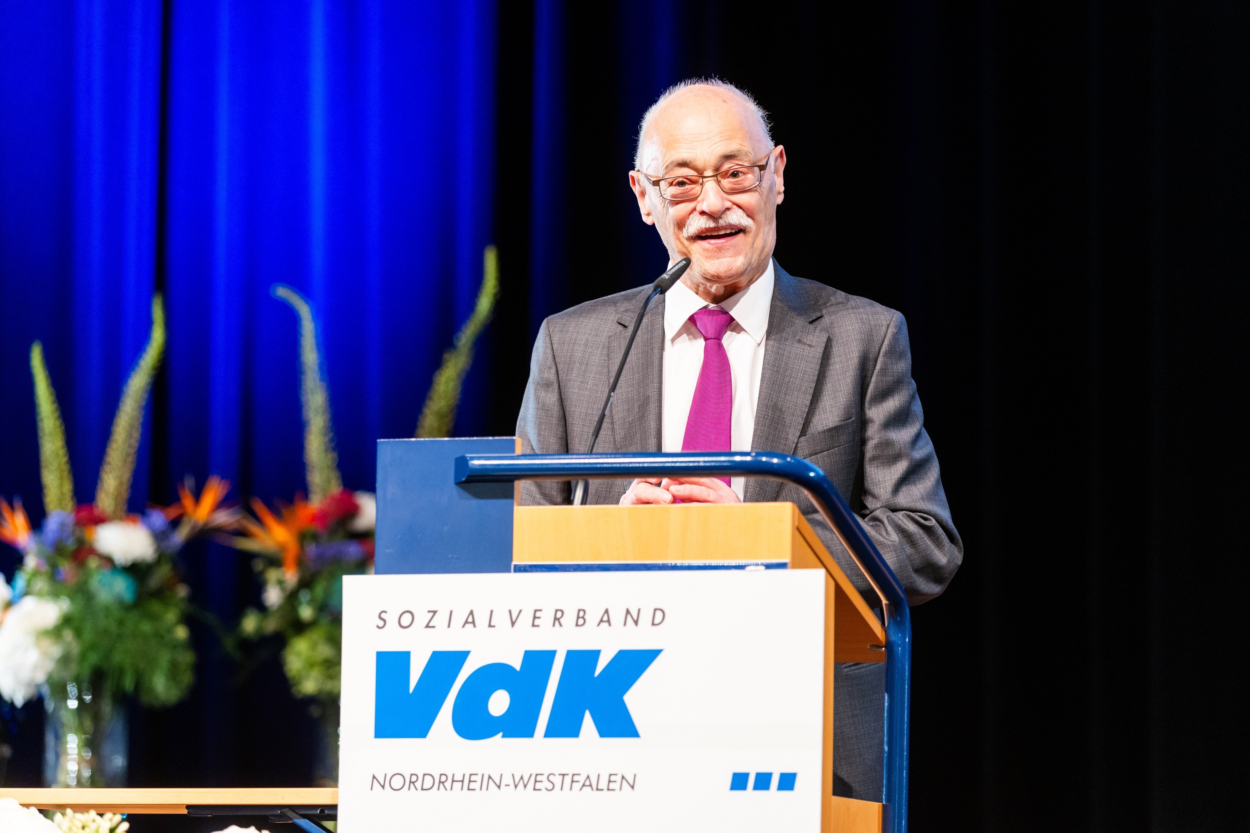 VdK-Landespräsident Horst Vöge steht in Anzug und mit lila Krawatte an einem Rednerpult mit VdK-Logo und spricht lächelnd ins Mikrofon. Im Hintergrund erkennt man Blumengestecke vor einem blauen Bühnenvorhang.