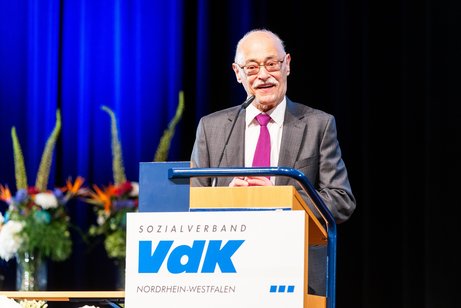 VdK-Landespräsident Horst Vöge steht in Anzug und mit lila Krawatte an einem Rednerpult mit VdK-Logo und spricht lächelnd ins Mikrofon. Im Hintergrund erkennt man Blumengestecke vor einem blauen Bühnenvorhang.