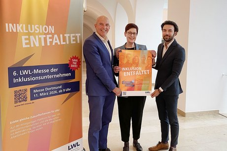 Drei Menschen stehen nebeneinander. Die Person in der Mitte h&auml;lt ein Plakat f&uuml;r die LWL-Messe der Inklusionsunternehmen in den H&auml;nden.