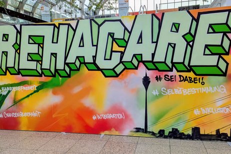 Bunte Graffitti-Wand mit den Buchstaben REHACARE und einer Skyline von Düsseldorf. 