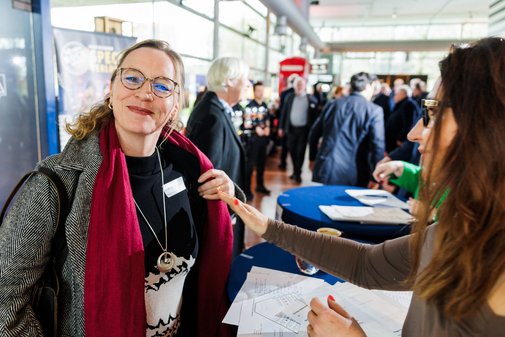 eine Frau mit Brille und rotem Schal zeigt stolz ihr Namensschild in die Kamera.