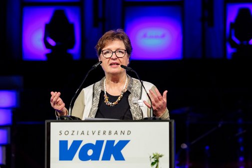 Prof. Dr. Ute Klammer steht an einem Pult auf der B&uuml;hne mit der Aufschrift VdK. Sie breitet beide Arme mit gestreckten H&auml;nden nach vorn aus. Sie tr&auml;gt eine Brille und hat etwas k&uuml;rzere, r&ouml;tliche Haare.