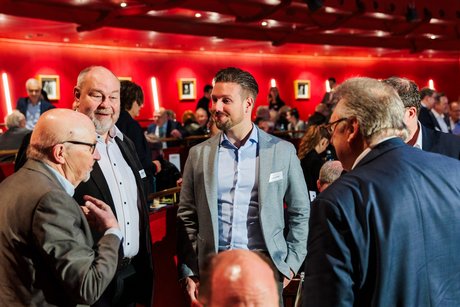 Vier Männer stehen redend beieinander. Im Hintergrund erkennt man einen Saal voller Menschen.