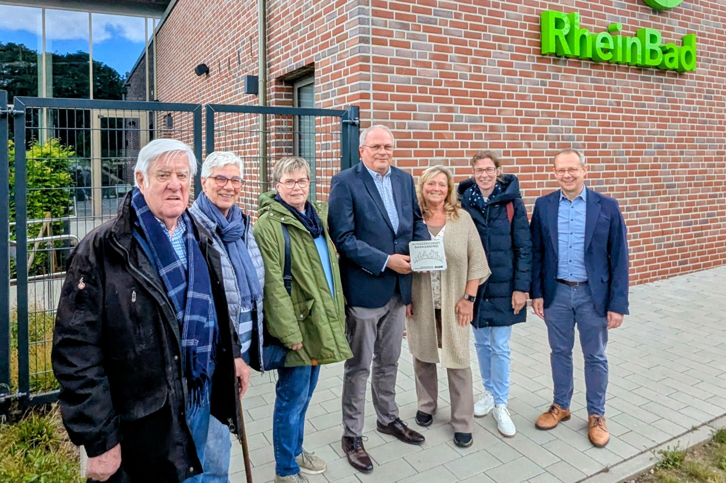 Sieben M&auml;nner und Frauen stehen vor einer roten Klinkerfassade mit gr&uuml;nem Logo RheinBad