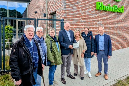 Sieben Männer und Frauen stehen vor einer roten Klinkerfassade mit grünem Logo RheinBad