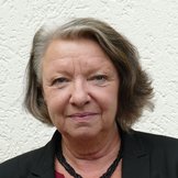 Porträt einer älteren Frau mit kinnlangen braungrauen Haaren in schwarzem Blazer mit Halskette vor einer weißen strukturierten Wand.