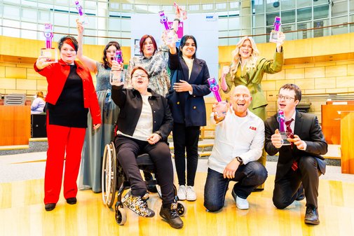 Zwei Männer und sechs Frauen, eine davon im Rollstuhl sitzend, haben sich für ein Gruppenfoto versammelt und halten jubelnd den pinken vilmA-Pokal in die Höhe. 