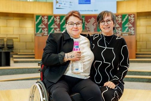 Eine Frau im Rollstuhl mit Brille und Kurzhaarschnitt hält den vilmA-Preis in der Hand. Neben ihr hockt eine Frau mit Brille und Kurzhaarschnitt in schwarzem Kleid. 