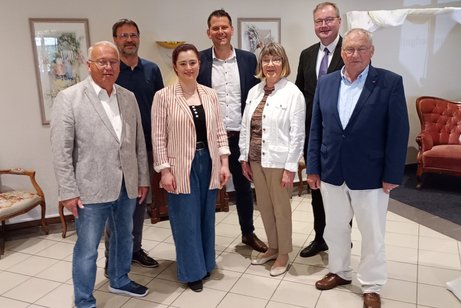 Eine Gruppe M&auml;nner und Frauen in Sakko, Blusen und Anz&uuml;gen steht in einem gefliesten Raum. Im Hintergrund ein roter Sessel und Gem&auml;lde an der wei&szlig;en Wand. 