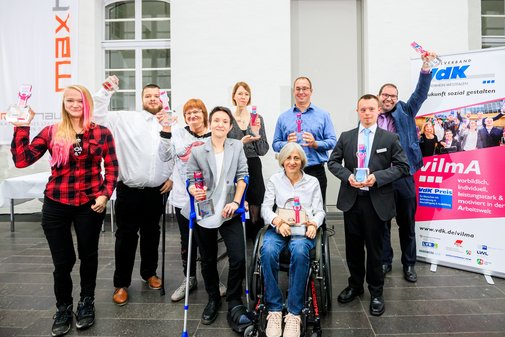 Eine Gruppe vilmA-Gewinner*innen, stehend oder im Rollstuhl sitzend, einige recken den Pokal in die Höhe. 