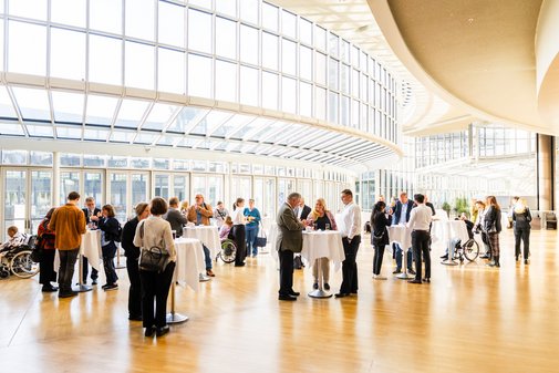 Menschen stehen an weißgedeckten Stehtischen in der lichtdurchfluteten Wandelhall des Düsseldorfer Landtag-Gebäudes.