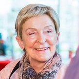 Portrait einer lächelnden älteren Frau mit blonden kurzen Haaren, rosa Bluse und geblümtem Halstuch.
