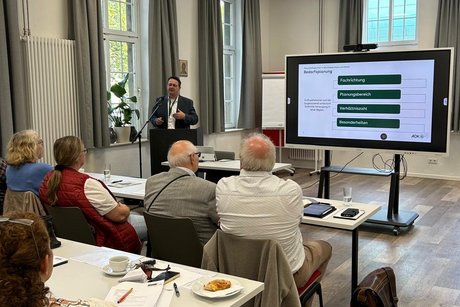 Blick über die vorderen Tischreihen im Seminarraum. Vorne ein Referent am Rednerpult mit Whiteboard-Präsentation neben sich.