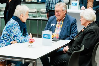 Gäste sitzen im Gespräch an einem Tisch. Auf dem Tisch eine Rose und eine Karte mit dem Aufdruck Soziales Forum.