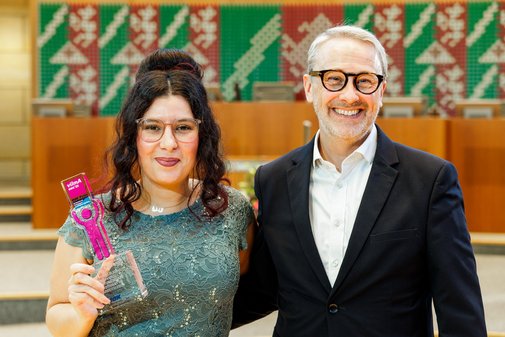 Eine Frau mit Brille und undklen, hochgesteckten Locken hält in einer Hand den vilmA-Preis. Neben ihr in Hemd und Anzug ein Mann mit Brille und grauem Kurzhaarschnitt. 