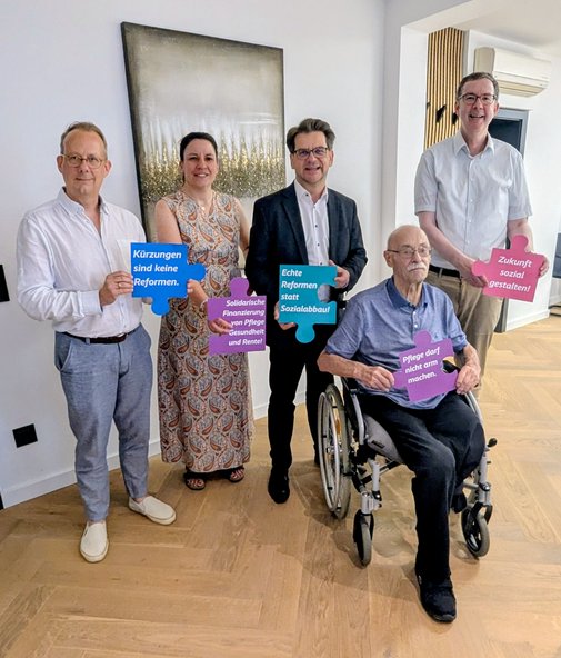 Fünf Menschen, davon einer im Rollstuhl, halten farbige Puzzleteile in den Händen, auf denen verschiedene Forderungen für einen gerechten Sozialstaat stehen.