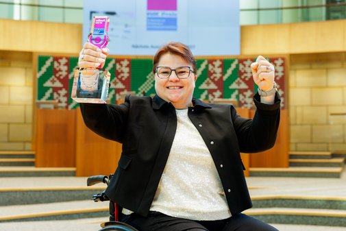 Eine Frau im Rollstuhl mit dunkler Kurzhaarfrisur und Brille h&auml;lt den vilmA-Preis in der linken Hand, die rechte ist ebenfalls emporgereckt zur Siegerfaust.