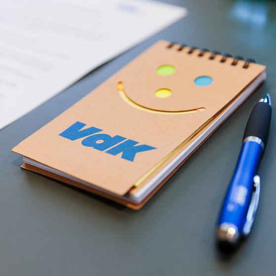 Ein kleiner Schreibblock mit ausgestanztem Lachgesicht und ein Kugelschreiber mit VdK-Logo liegen auf einer dunklen Tischplatte