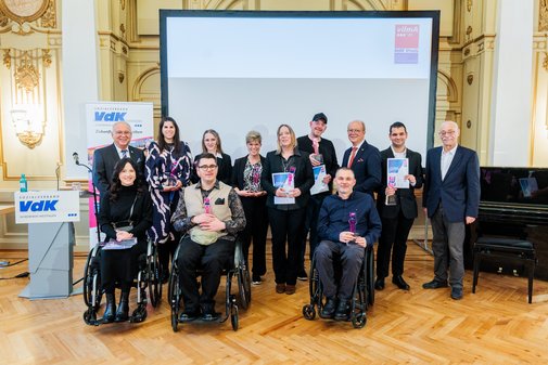 Gruppenfoto der vilmA-Preisträger*innen und Laudator*innen vor einer Presseleinwand, stehend und im Rollstuhl sitzend. 