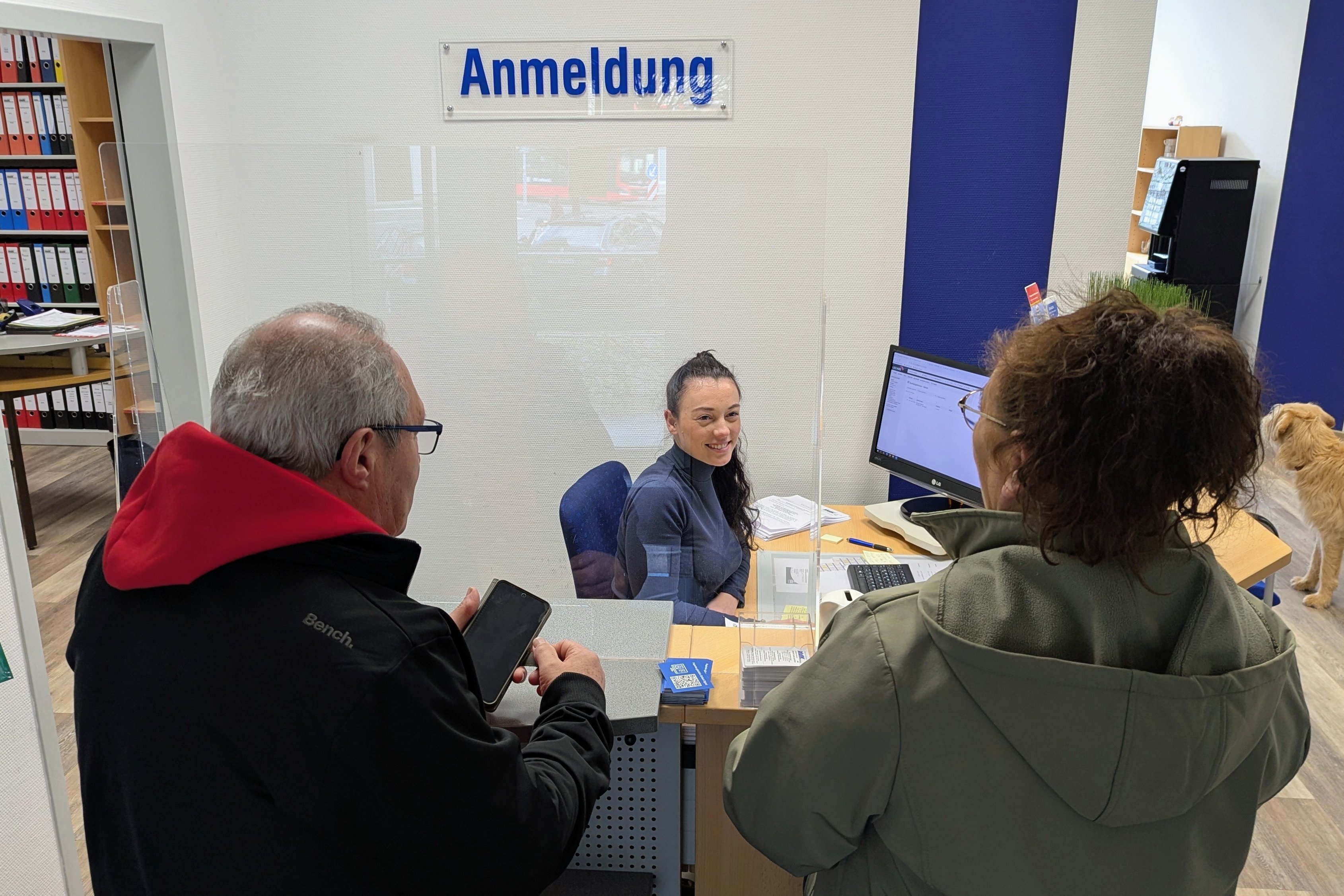 Zwei Menschen stehen an einer Empfangstheke, dahinter sitzt löchelnd eine junge Frau mit dunklen Locken an einem PC. Über ihr in blauer Schrift ein Schild Anmeldung.