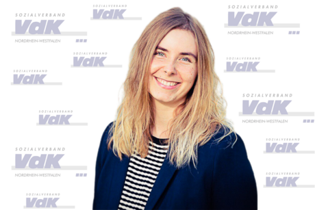 Eine lächelnde Person mit langen, blonden Haaren trägt ein schwarz-weiß gestreiftes Shirt und einen dunkelblauen Blazer vor einem Hintergrund mit mehreren "Sozialverband VdK Nordrhein-Westfalen" Logos.