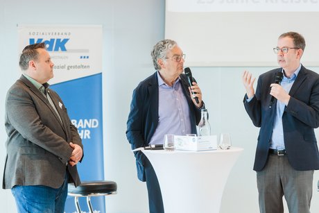 Drei Männer in Anzug stehen nebeneinander um einen Stehtisch. Im Hintergrund ein Roll-Up mit Logo des VdK NRW.
