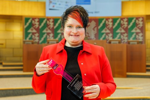 Eine Frau mit rotem Jacket und dunklem Kurzhaarschnitt mit roter Ponystr&auml;ne h&auml;lt l&auml;chelnd den vilmA-Preis in den H&auml;nden.